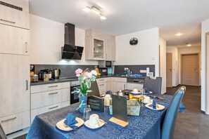 A modern kitchen with light wood cabinets, a microwave, and a dining area set for two with a blue tablecloth and a vase of flowers.