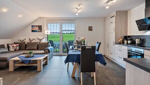 A modern living room with a sofa, coffee table, dining table, and chairs. There is a kitchen area with cabinets and a stove. A large sliding glass door leads to a grassy outdoor area. - Apartment 'Alpenpanorama, 76qm, 2 Schlafräume' with Mountain View, Shared Terrace and Wi-Fi (Höchenschwand)