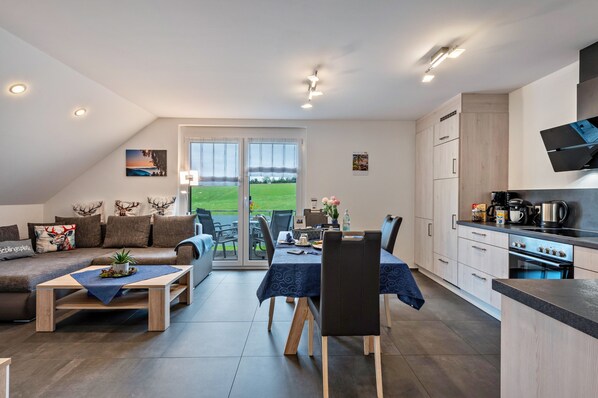 A modern living room with a sofa, coffee table, dining table, and chairs. There is a kitchen area with cabinets and a stove. A large sliding glass door leads to a grassy outdoor area.
