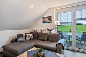A modern living room with a sectional sofa, a small table with a plant, and a large sliding glass door leading to an outdoor area with a table and chairs.