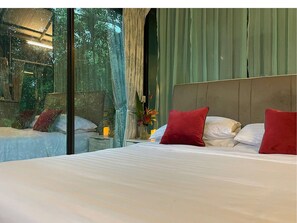 A bedroom with a large bed, a nightstand, a vase with flowers, and a view of greenery through a window. - Villa Sri Kampung  (Langkawi)