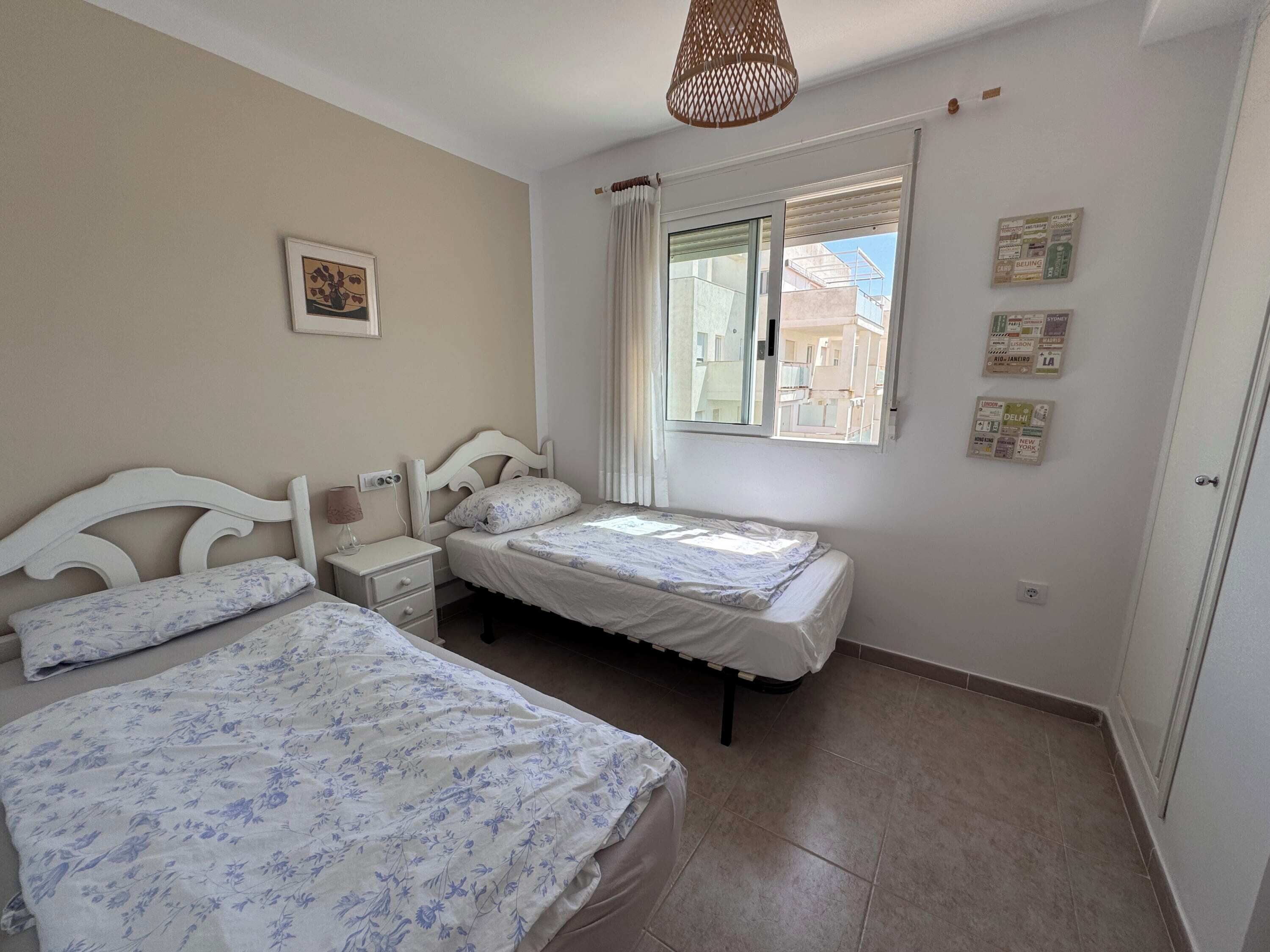 A bedroom with two beds, a nightstand, a window with a view of buildings, and a hanging light fixture.