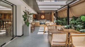 Un café moderne doté d’un grand luminaire circulaire au centre, de tables et de chaises en bois, et de grandes plantes en pot.