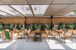 Un vaste espace intérieur doté d’une table et de chaises en bois, entouré de grandes plantes en pot, et d’un plafond haut agrémenté de lucarnes.