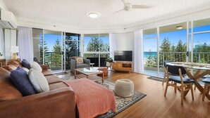 Amplia sala de estar con sofá, mesa de centro y televisor, ventanas grandes con vistas a árboles y un balcón.