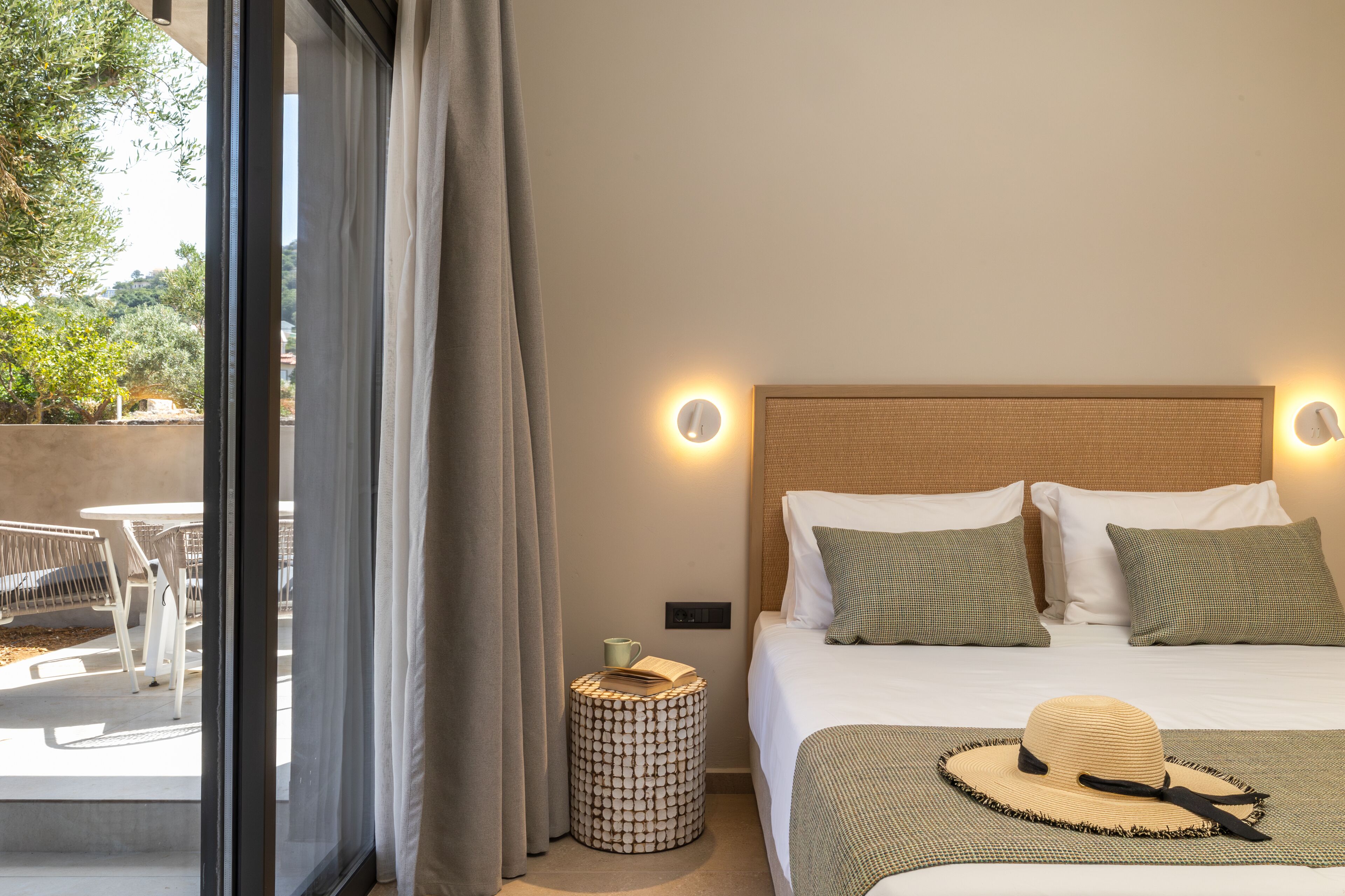 Une chambre d’hôtel avec un lit, des lampes de chevet, une petite table ronde avec un livre et une tasse, et une vue sur l’extérieur par une fenêtre.