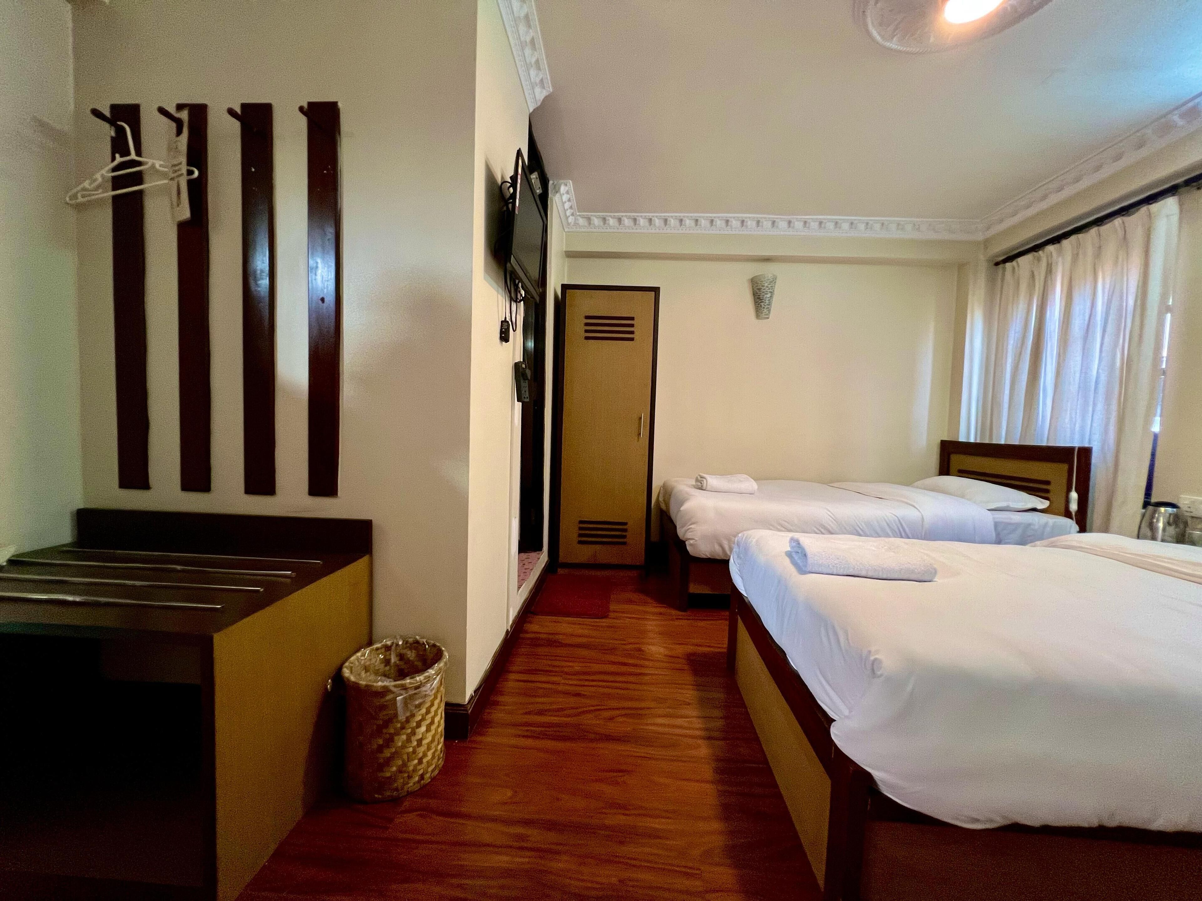 A hotel room with two beds, a wooden bench, a waste bin, and a coat rack with hangers.