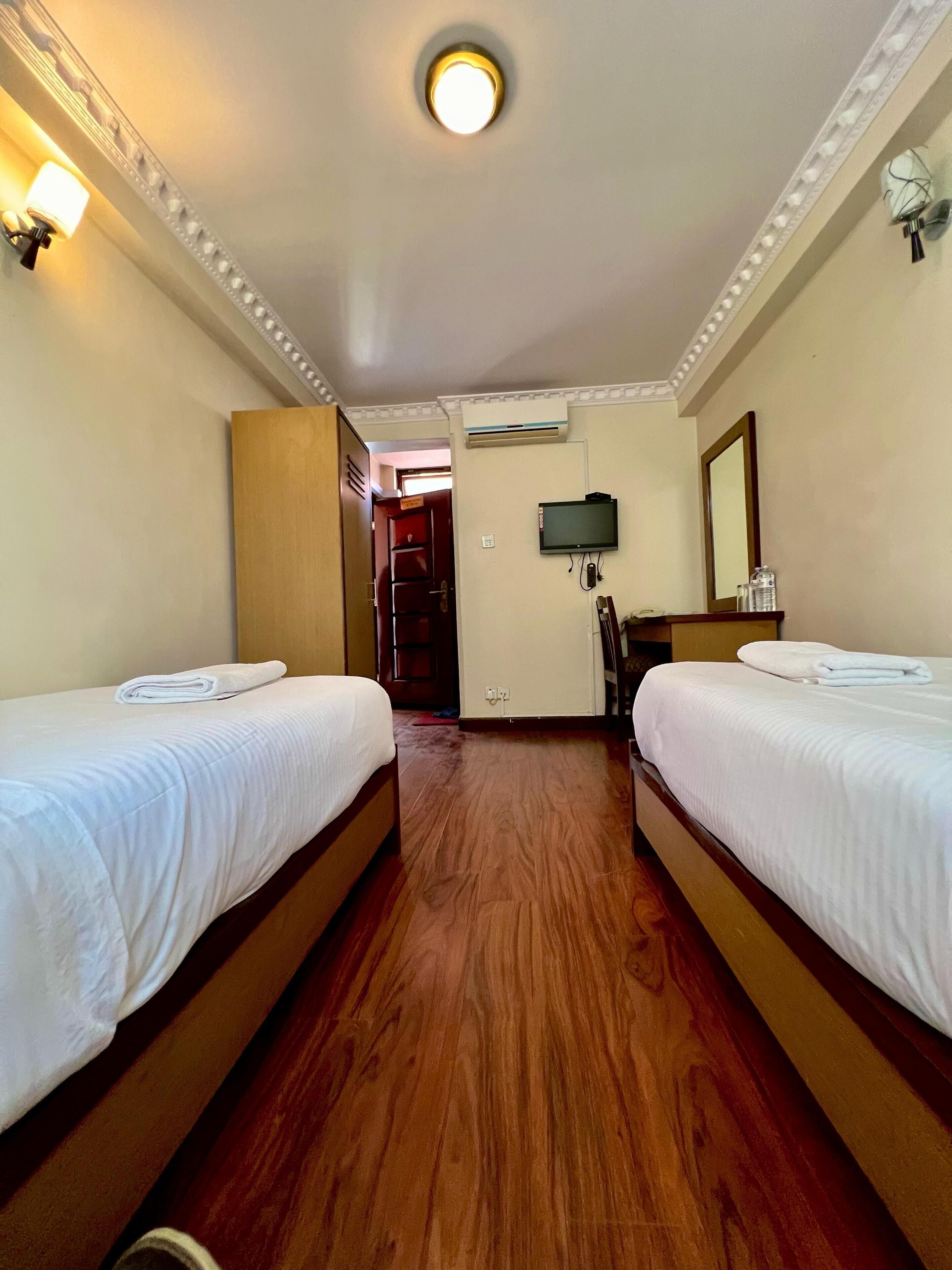 A hotel room with two beds, wooden flooring, a television, and an air conditioning unit.