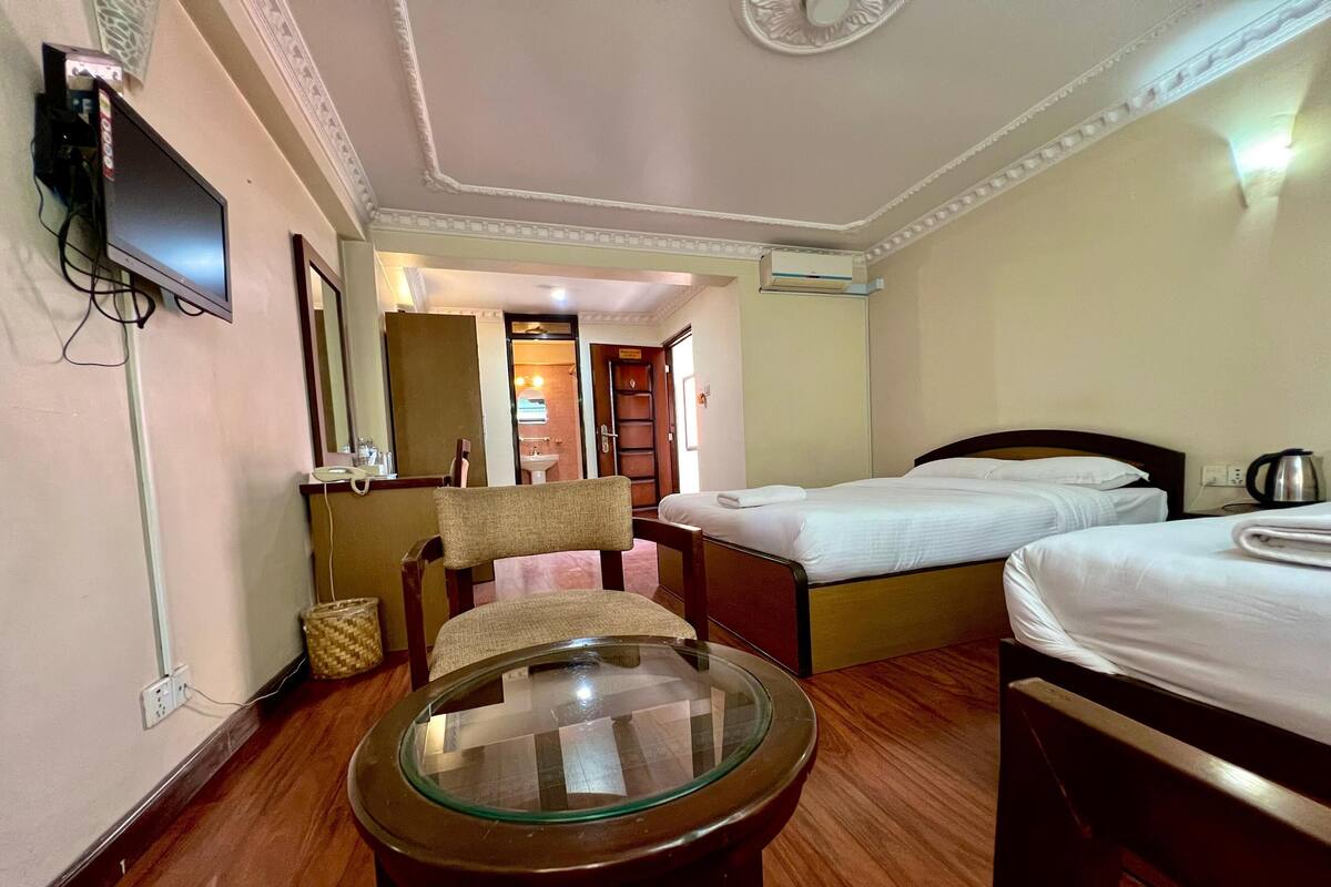 A hotel room with two beds, a small table, a chair, a television, and a bathroom visible through an open door.