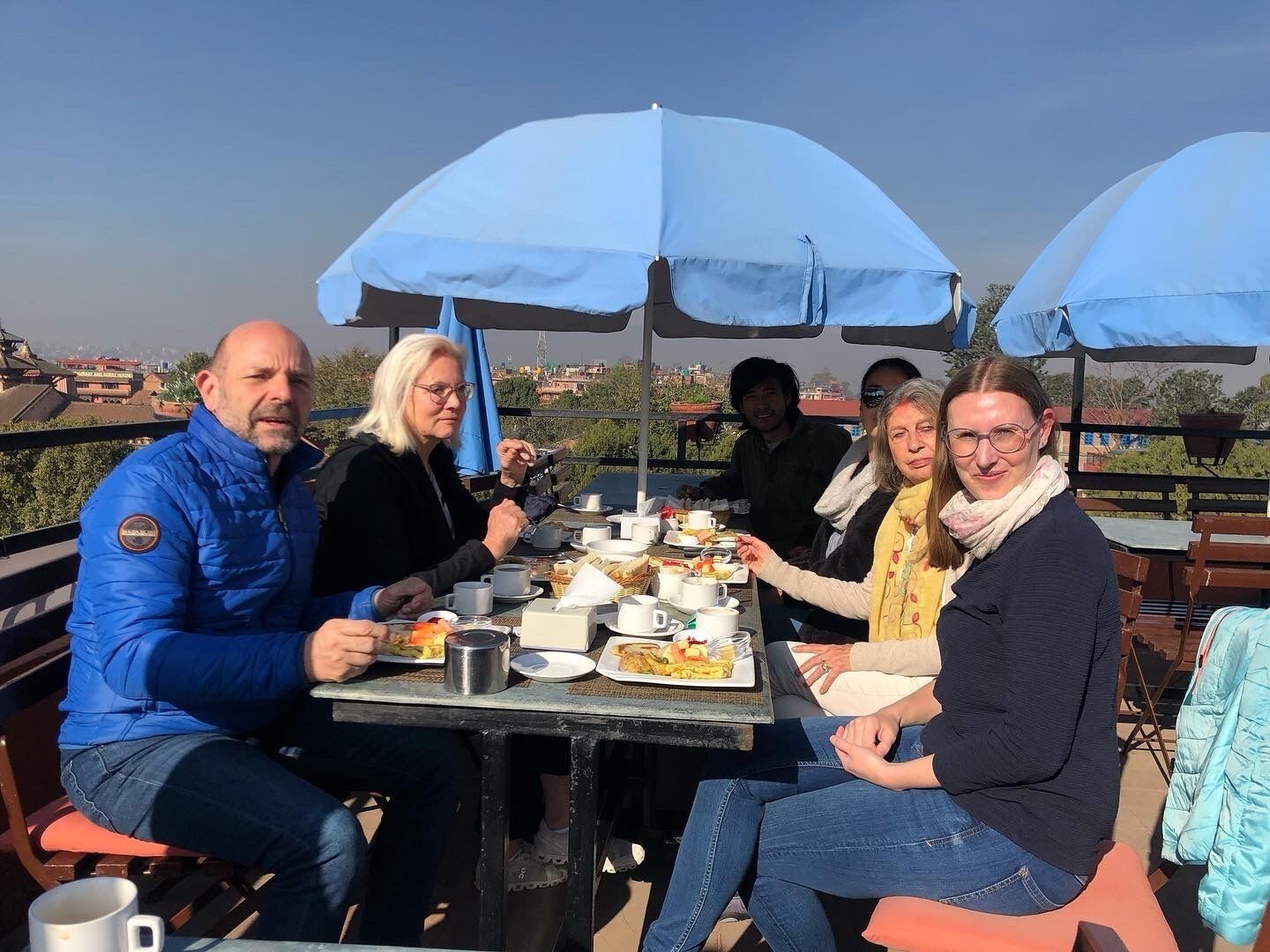 A group of people dining outdoors at a table with blue umbrellas, surrounded by a scenic view.