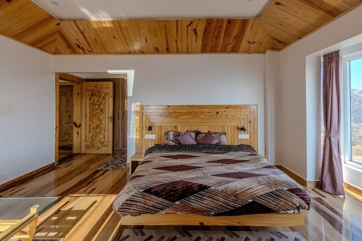 A bedroom with a wooden headboard, a large bed with patterned bedding, a glass coffee table, and a view of the outdoors through a window.