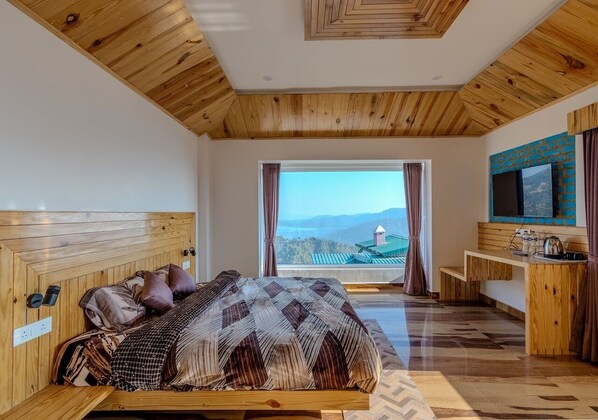 Un dormitorio con techo de madera, una cama grande con ropa de cama estampada, vistas a montañas a través de una ventana y una pequeña área de kitchenette.