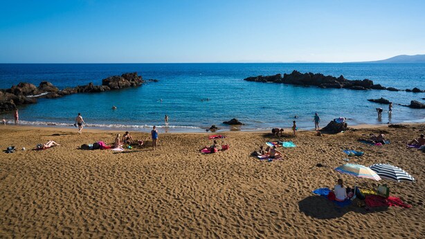 Una playa con gente tomando el sol y nadando, afloramientos rocosos en el agua, y un cielo azul despejado.
