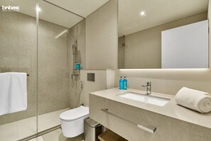 A modern bathroom with a glass shower enclosure, a white toilet, a sink with a mirror, and a towel on the counter. - bnbme | La Vie | 1 BDR (Dubai)