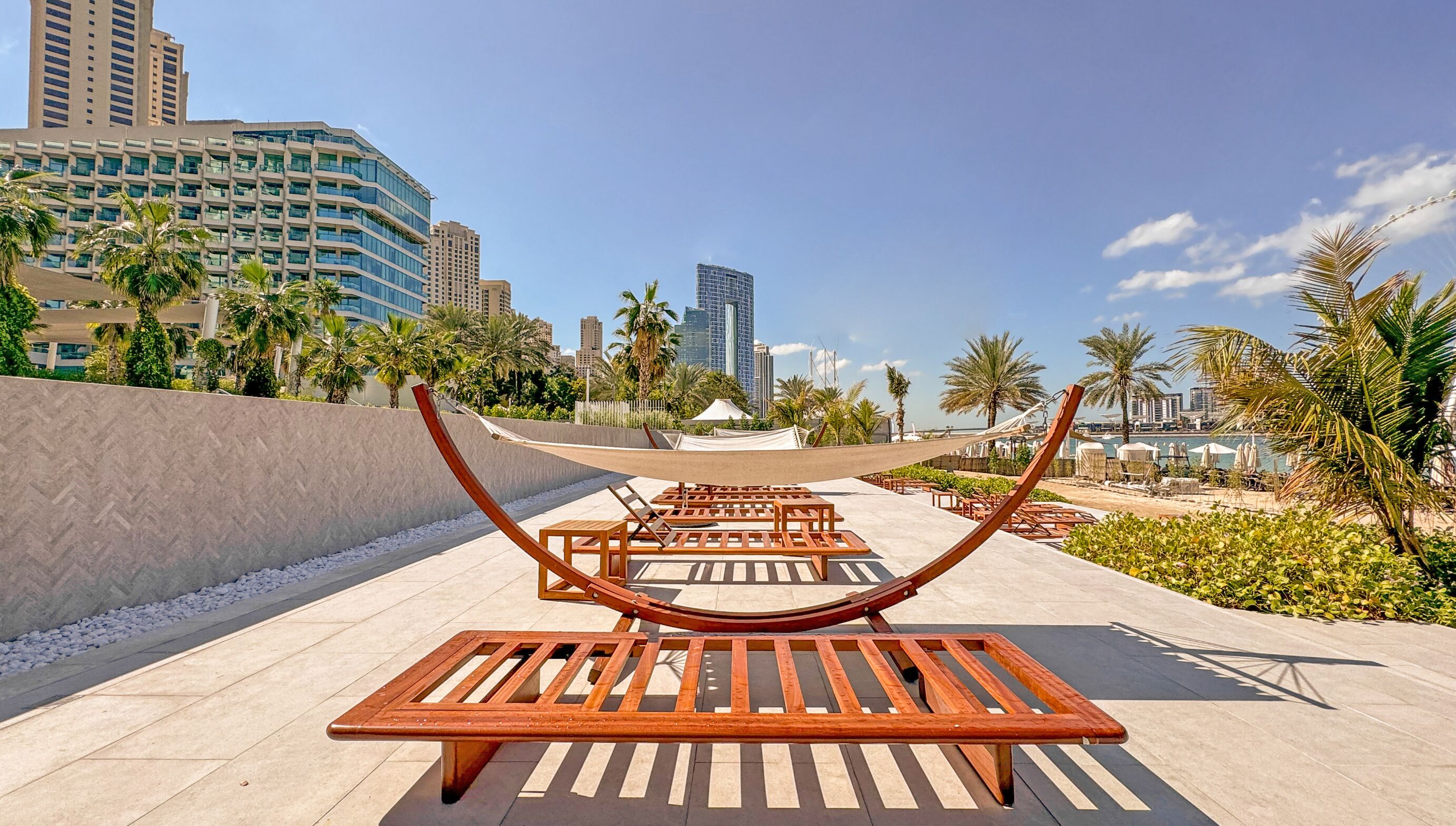 A row of wooden lounge chairs and tables in an outdoor setting with palm trees and a cityscape in the background.
