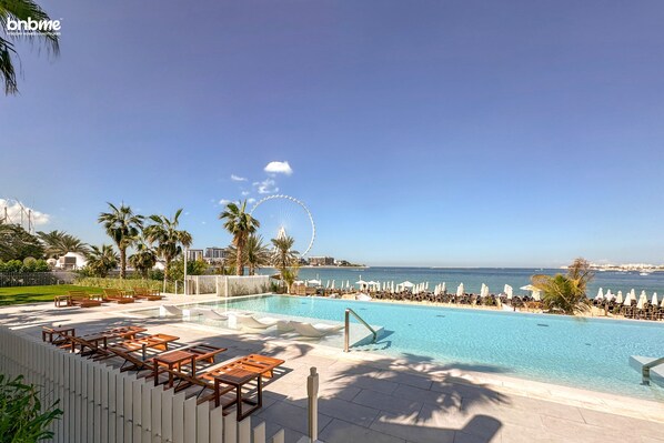 A poolside area with lounge chairs and umbrellas, a clear sky, and a distant Ferris wheel. - bnbme | La Vie | 1 BDR (Dubai)