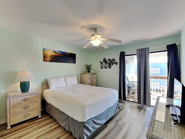 A hotel room with a bed, a nightstand, a lamp, a painting, a plant, a ceiling fan, and a balcony with curtains.