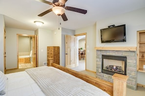 A bedroom with a large bed, wooden headboard, a ceiling fan, a flat-screen TV mounted above a stone fireplace, and a wooden door leading to a bathroom. - Nisswa Townhome w/ Boat Slip + Gull Lake Access! (Nisswa)