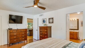 A bedroom with a bed, wooden dresser, TV, ceiling fan, and a bathroom visible through an open door. - Condo in PGA National Resort w/ Pool & Spa! (Palm Beach Gardens)