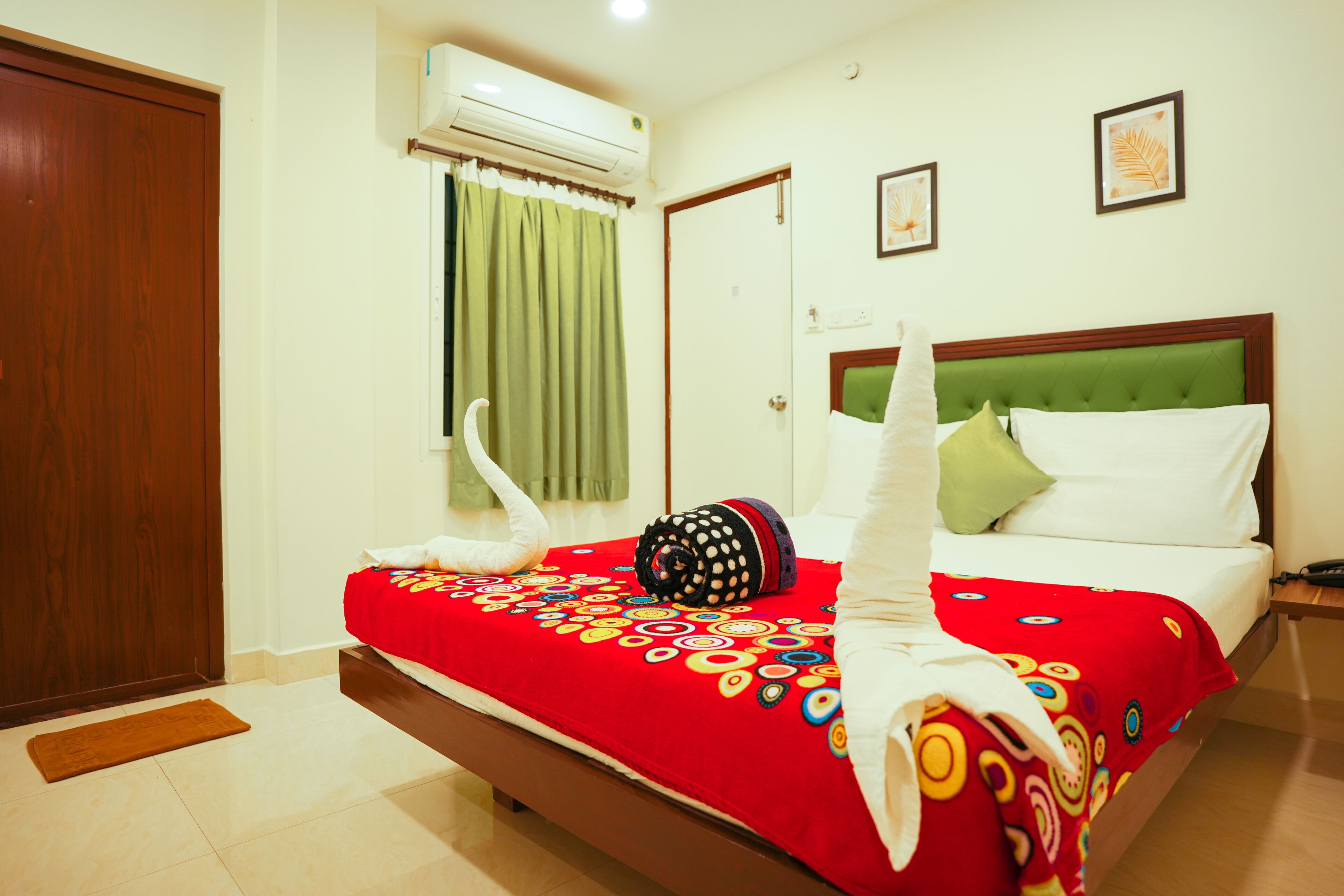 A hotel room with a bed, a wooden door, an air conditioning unit, a green curtain, and a wall with two framed pictures.