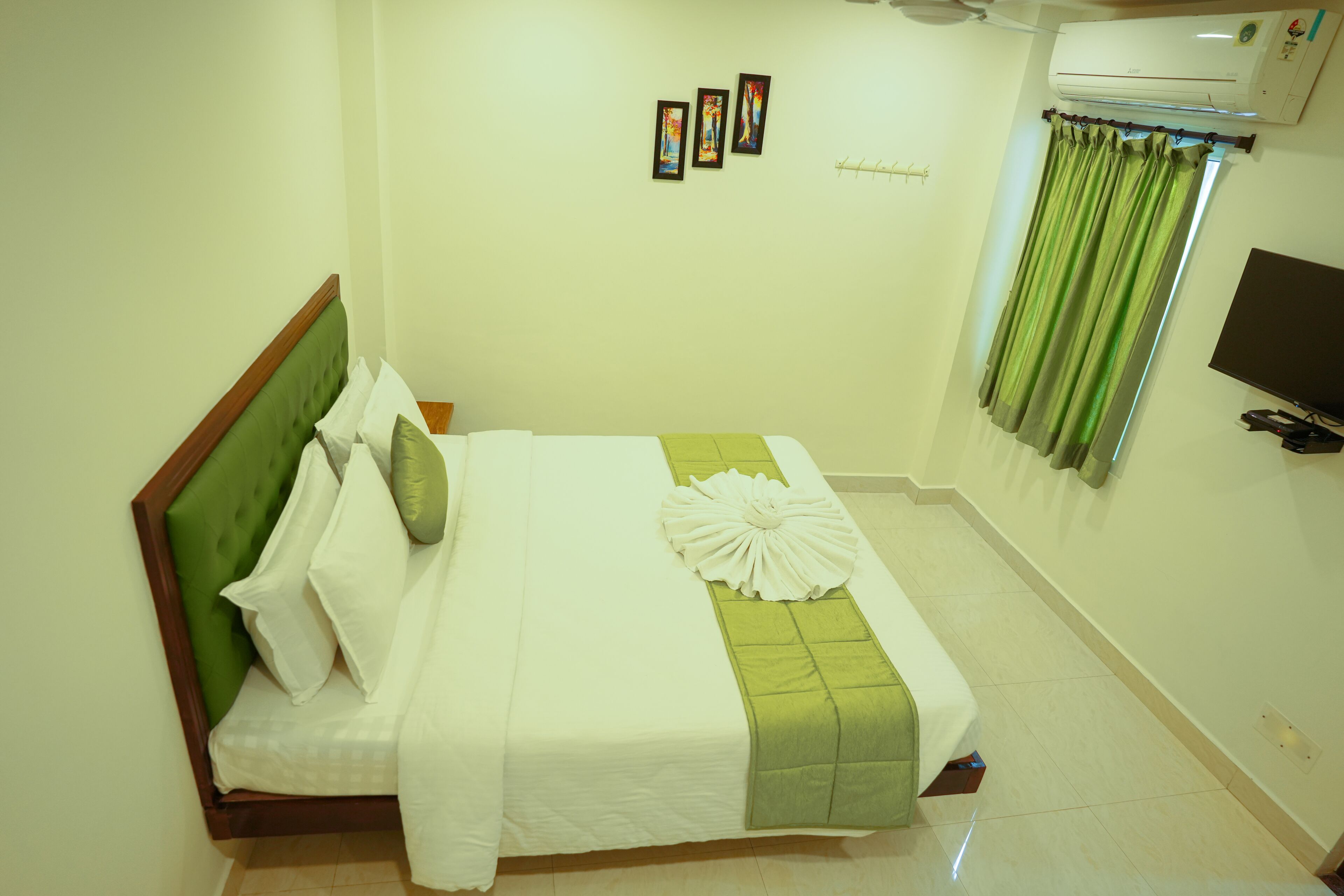 Una cama bien tendida con cabecera verde y ropa de cama blanca, almohada verde y blanca a rayas y una almohada decorativa blanca. Hay tres cuadros enmarcados en la pared, una cortina verde y un televisor de pantalla plana montado en la pared.