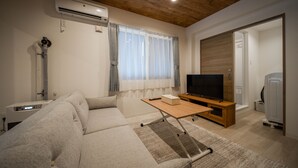 A modern living room with a wooden ceiling, a large window with curtains, a flat-screen TV on a wooden stand, a sofa, and a small table. - Astoria105 (Asahikawa)