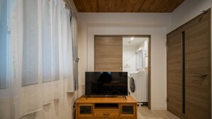 A modern living room with a flat-screen TV on a wooden stand, a wooden dining table, and a kitchen area visible through an open door. - Astoria105 (Asahikawa)