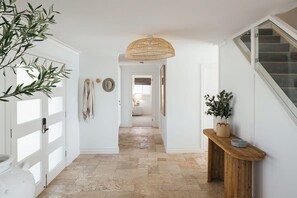 A hallway with a stone floor, white walls, a wooden console table with a potted plant, and a woven ceiling light.