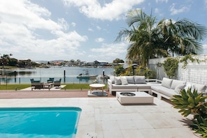 A pool area with a white sofa and a table, surrounded by palm trees and a view of a lake with boats.