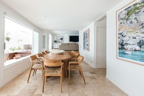 A spacious dining area with a round wooden table and chairs, a sofa in the background, and a large framed picture of a rocky landscape.
