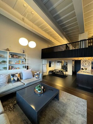A spacious living area with a modern loft design, featuring a high ceiling, a mezzanine level, and a large black coffee table.