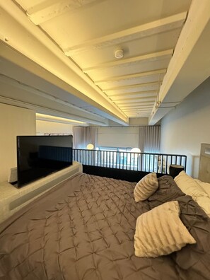 A bedroom with a bed, a flat-screen TV, a balcony with curtains, and a view of the outside.