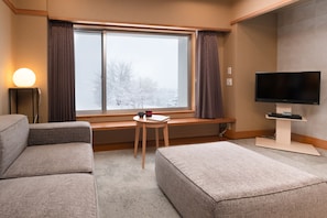 Et hotelværelse med et stort vindue, et fladskærms-tv, en sofa, et lille bord med en kop og en bog, og en lampe.