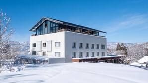 A modern two-story building with large windows and a flat roof, surrounded by snow-covered landscape and mountains. - Hokuryuko Hotel North Nagano (Iiyama)