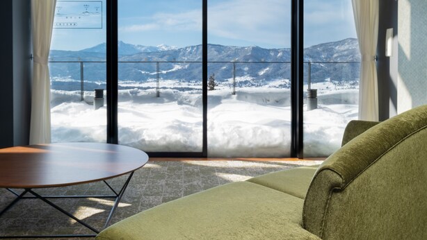 A room with a green sofa, a small round table, and a large window offering a view of snow-covered mountains. - Hokuryuko Hotel North Nagano (Iiyama)
