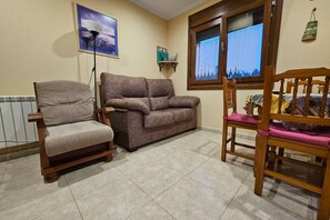 A living room with a sofa, two armchairs, a wooden chair, a radiator, a window with curtains, and a wall-mounted shelf. - 30A01 Apartamentos rurales La Brañela (Castropol)