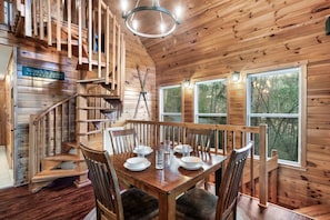 A wooden cabin interior with a dining table set for four, a spiral staircase, and large windows offering a view of the forest. - 2 BED|2.5 BATH-Butterfly Views-Close to Dollywood! (Sevierville)