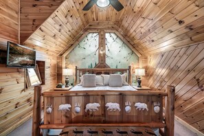 A wooden cabin bedroom with a sloped ceiling, a bed with a wooden headboard featuring animal carvings, two bedside lamps, and a television on the wall. - 2 BED|2.5 BATH-Butterfly Views-Close to Dollywood! (Sevierville)
