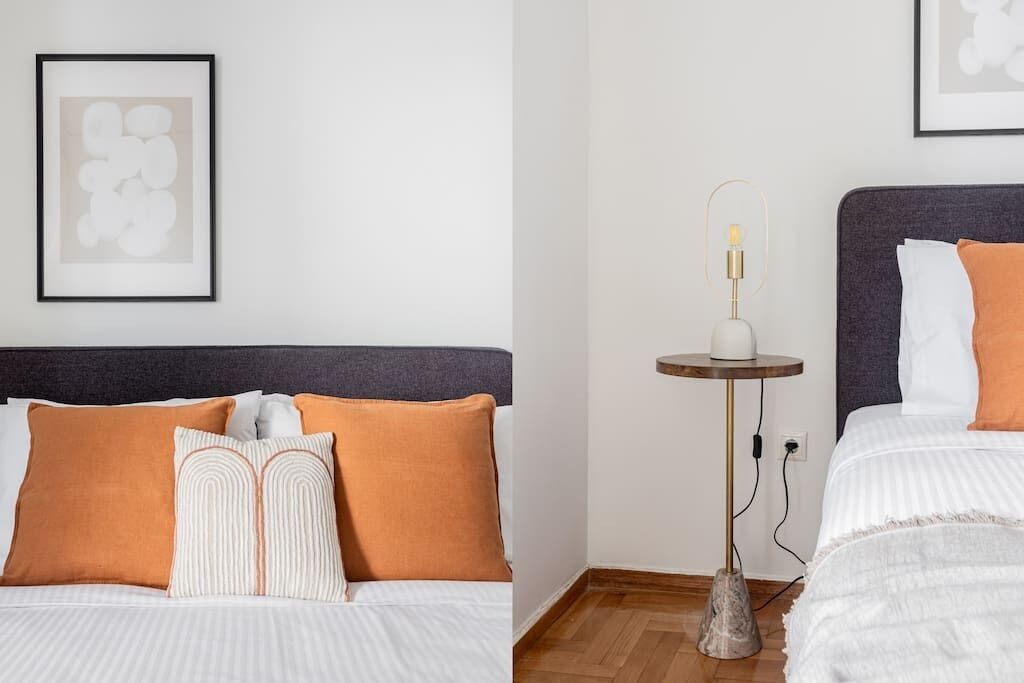 Una cama bien hecha con cabecera gris, ropa de cama blanca y dos almohadas naranjas. Un cuadro enmarcado cuelga de la pared sobre la cama.