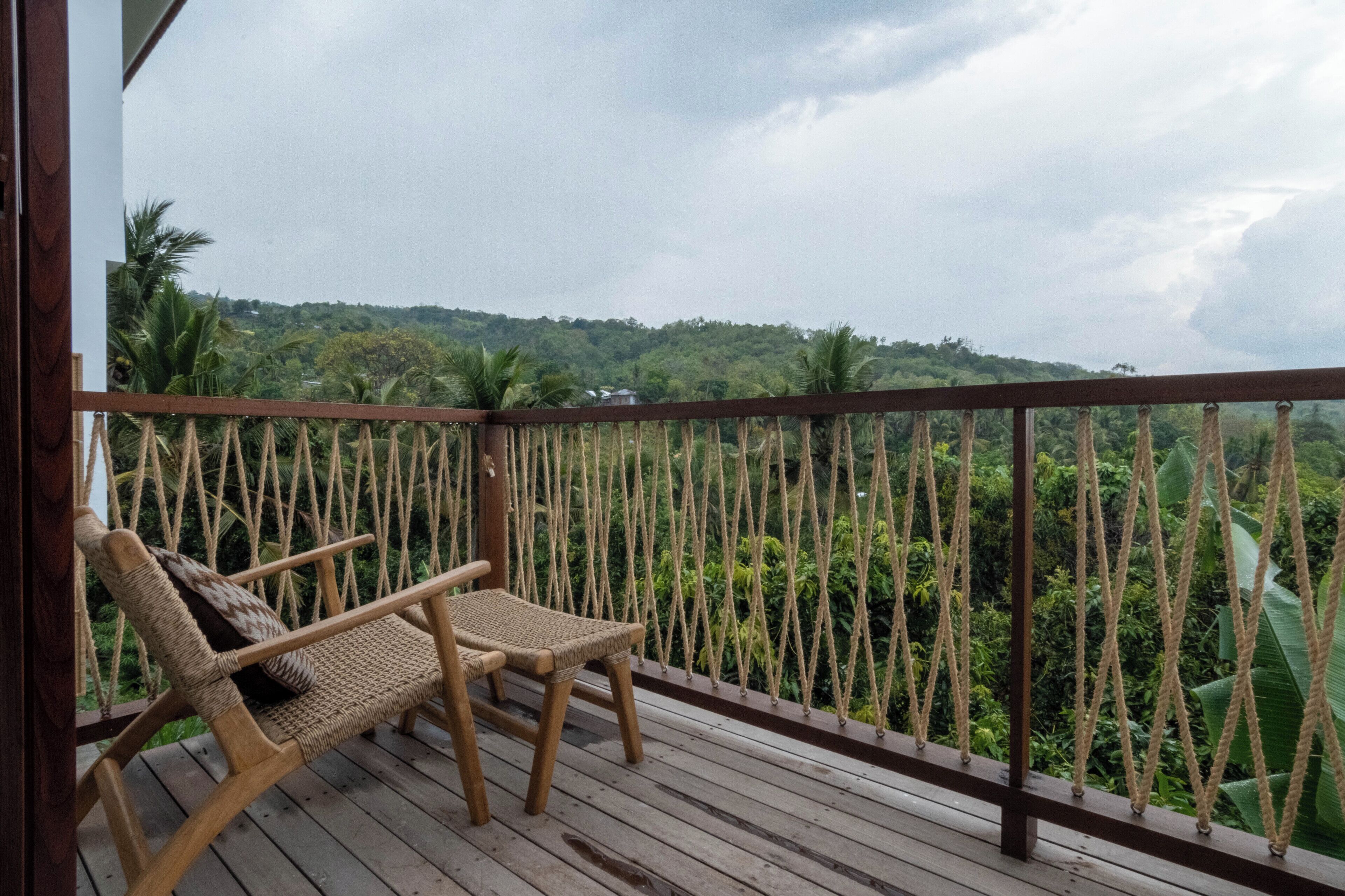 Una terraza de madera con dos sillones de mimbre y una barandilla de cuerda, con vistas a un paisaje verde exuberante.