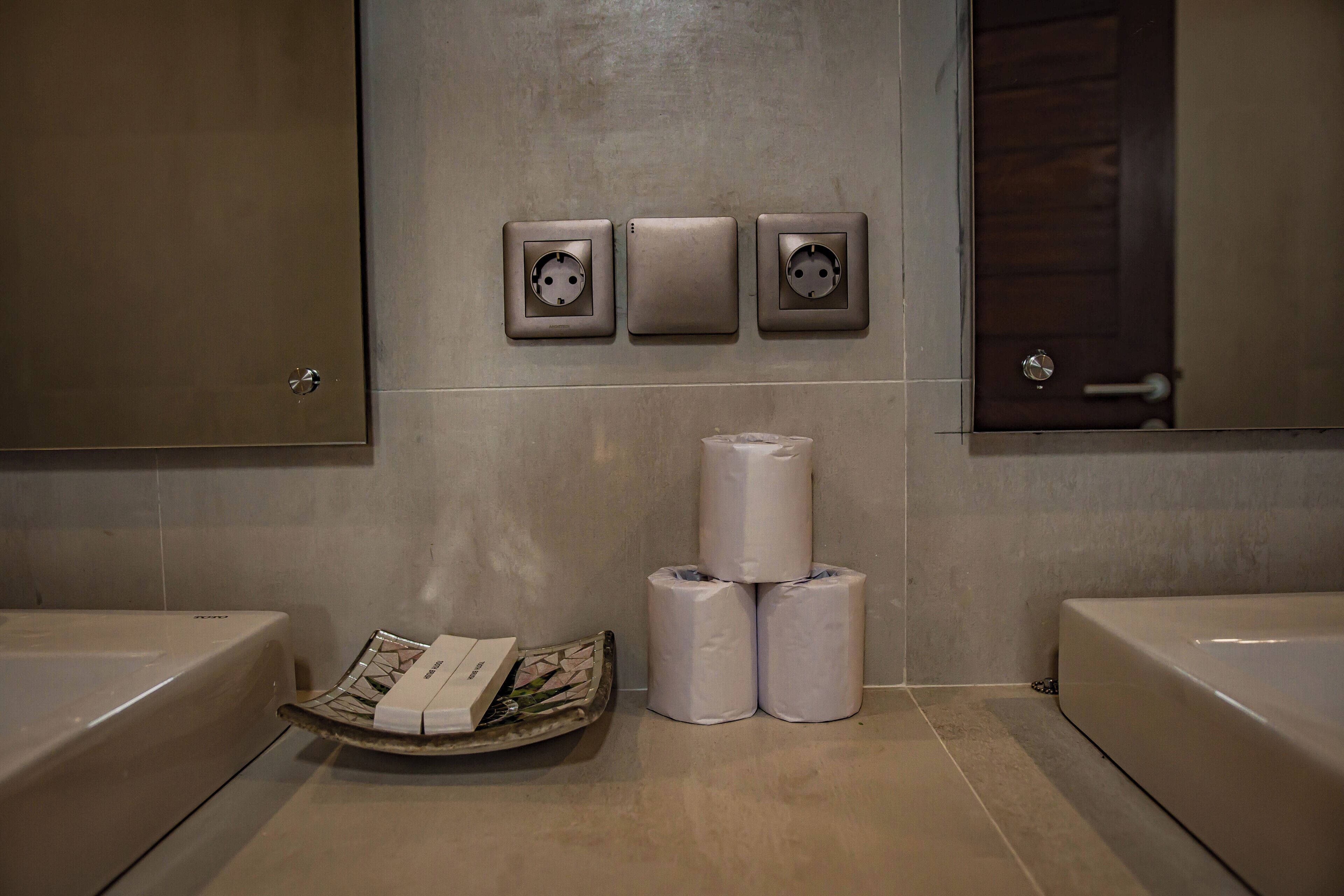 A bathroom with two sinks, three electrical outlets on the wall, and a stack of toilet paper.