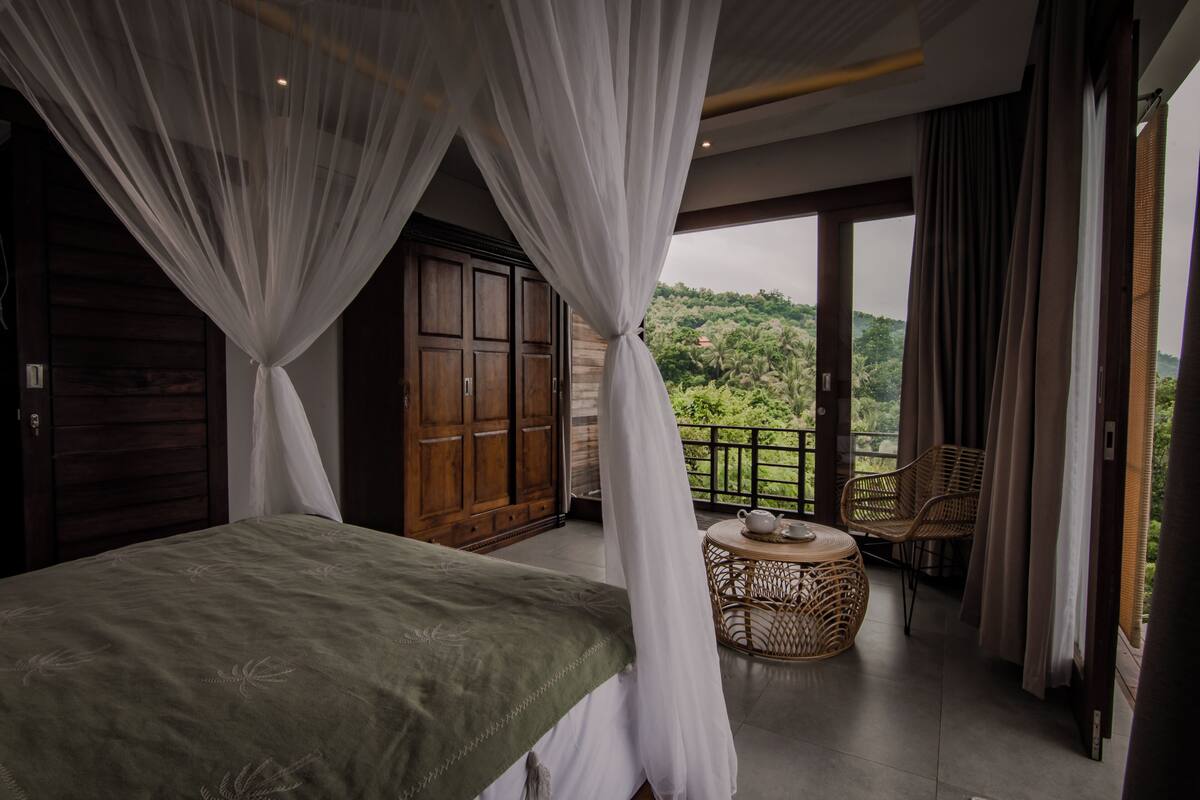 Ein Schlafzimmer mit Baldachinbett, einem Balkon mit Korbmöbeln und Blick auf Grünflächen.