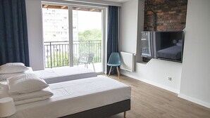A modern bedroom with a large bed, a TV mounted on the wall, and a balcony with a view of buildings and greenery. - MoHo Modern Hostel (Wroclaw)