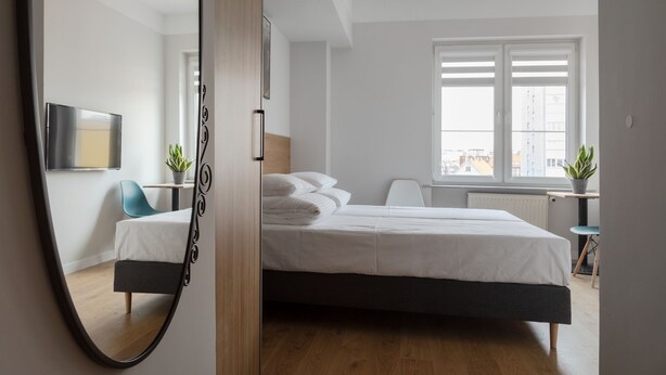 A modern bedroom with a bed, a mirror, a TV, and a window with a view of buildings. - MoHo Modern Hostel (Wroclaw)