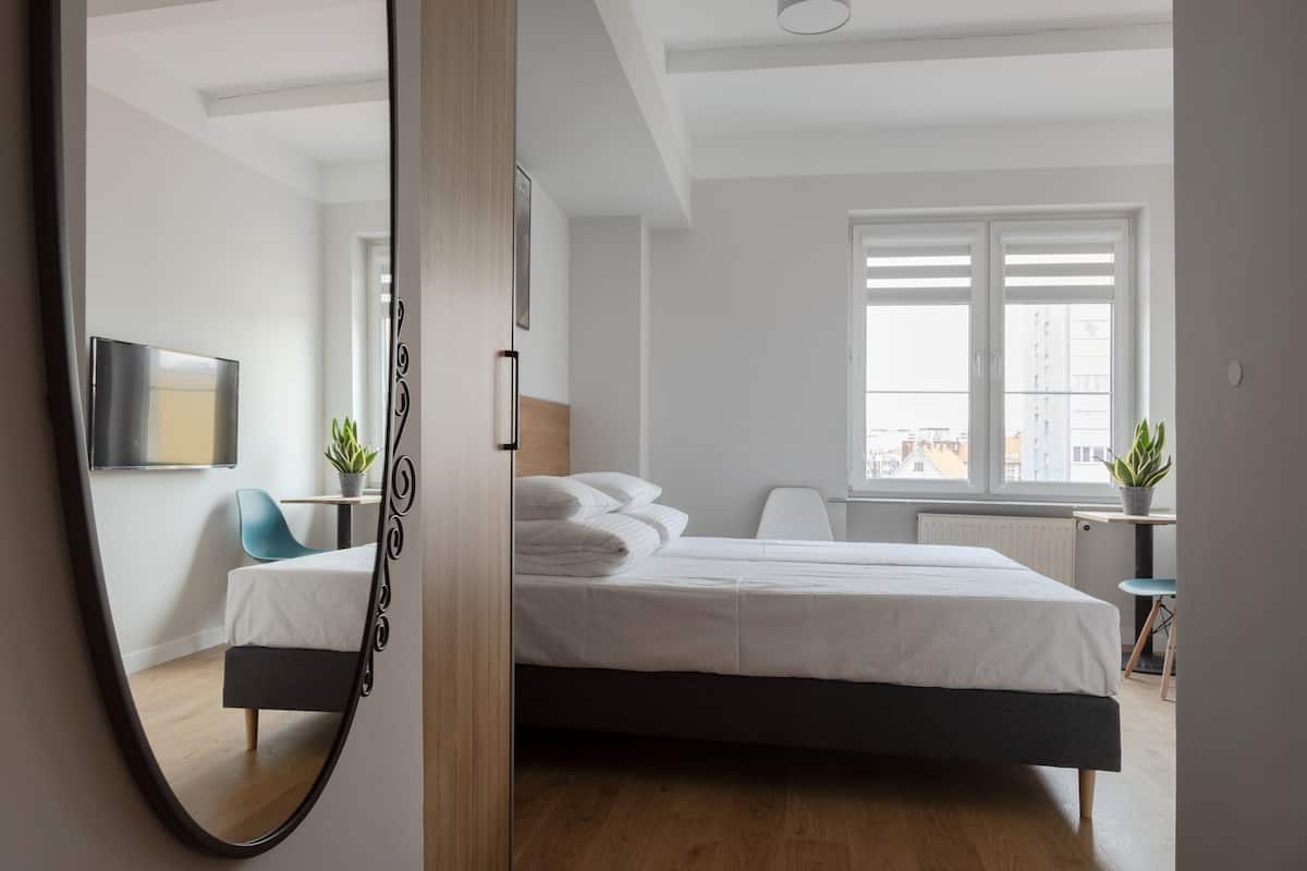 A modern bedroom with a bed, a mirror, a TV, and a window with a view of buildings.
