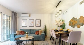 Una sala moderna con sofá, mesa de centro, mesa de comedor y sillas. Hay obras de arte enmarcadas en la pared, una planta en maceta y una ventana con cortinas.