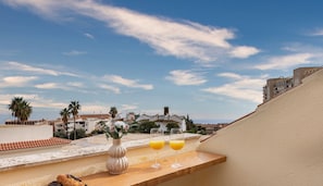A rooftop terrace with a table set for breakfast, including pastries and orange juice glasses, overlooking a residential area with palm trees and buildings.
