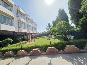 Um edifício moderno com vários andares e grandes janelas. Possui um jardim bem cuidado, com grama, arbustos e algumas árvores.