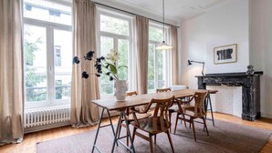 A dining area with a wooden table and chairs, a vase with flowers, a fireplace, and large windows with curtains. - The Handshake House by Shway (Antwerpen)
