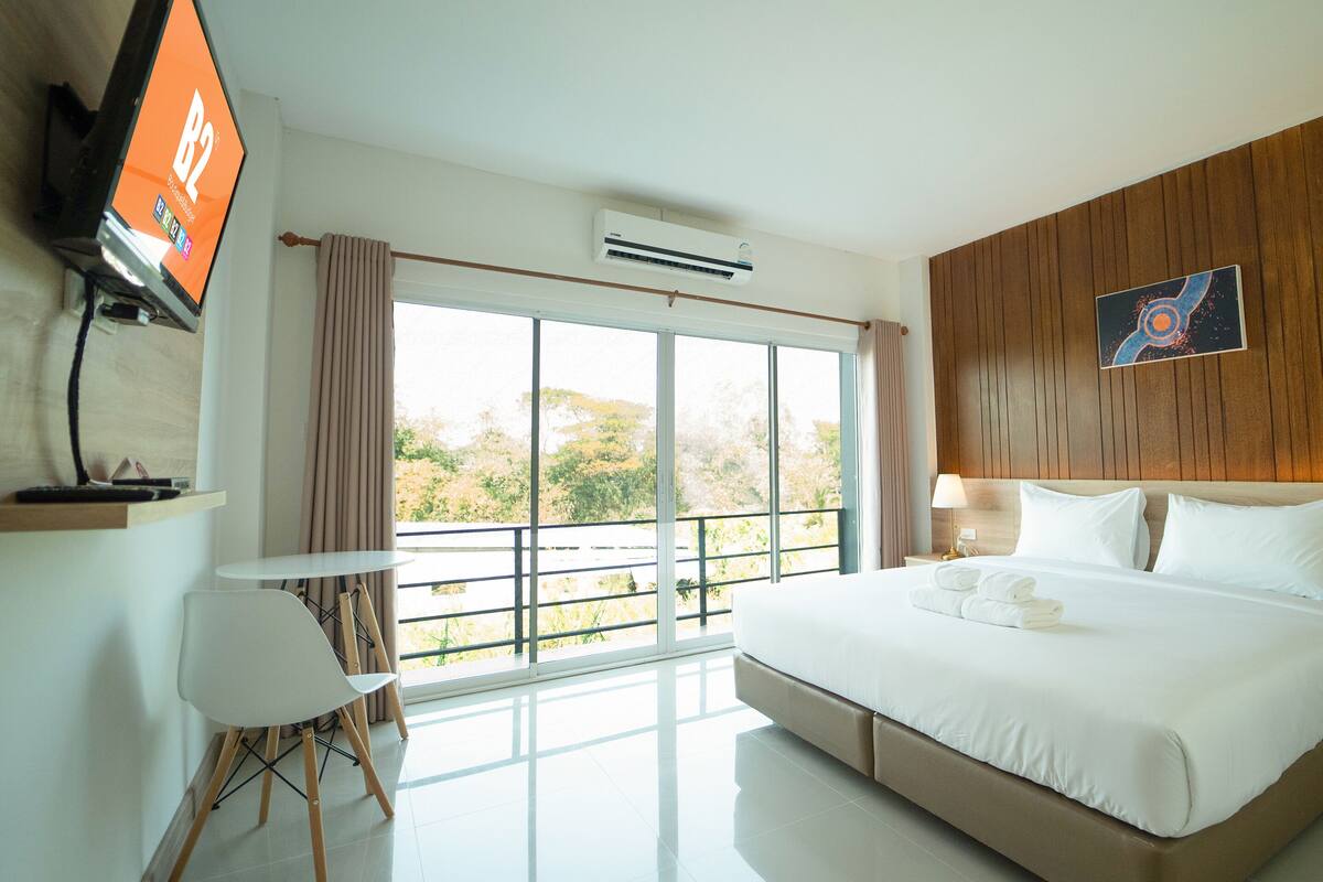 A hotel room with a large bed, a desk with a chair, a TV, and a window with a view of trees.